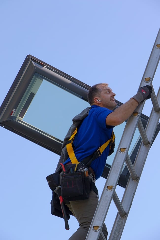 A skylight installer carrying a skylight up a ladder