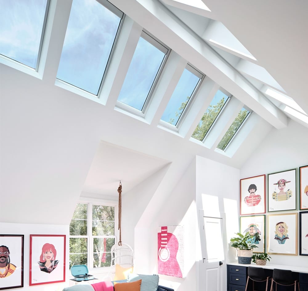 Roof ridge skylights with shades brighten a living room with blue and pink decor and portrait art on two walls.