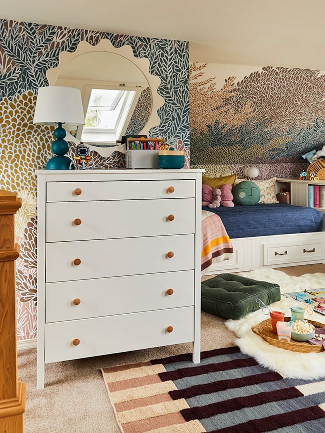 A white chest of drawers in a colorful childrens room with a sun shaped mirror reflecting a skylight on the opposite wall