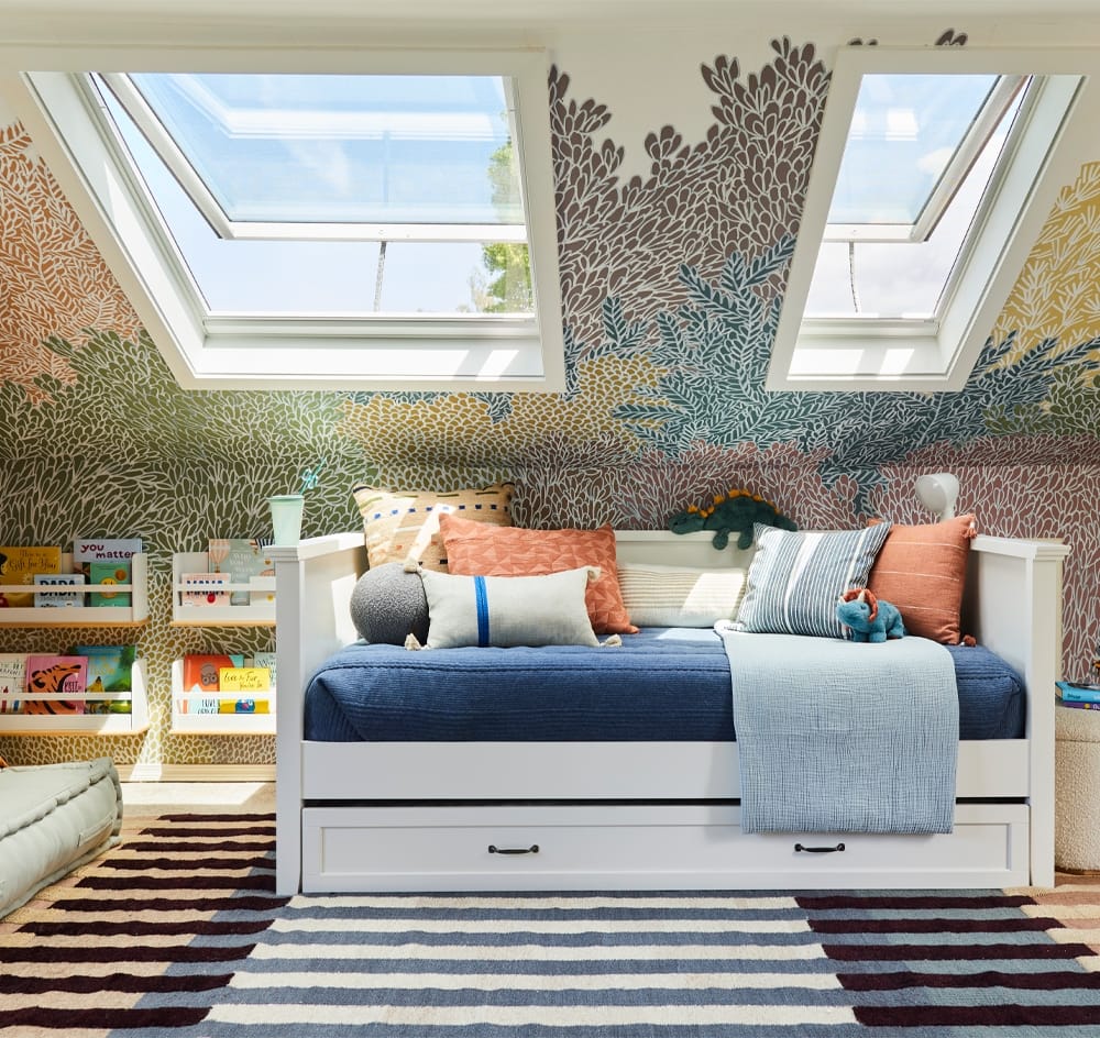 A children's bedroom with two skylights and colorful tree-themed wallpaper and a trundle bed flanked by a blue, maroon and white striped rug.