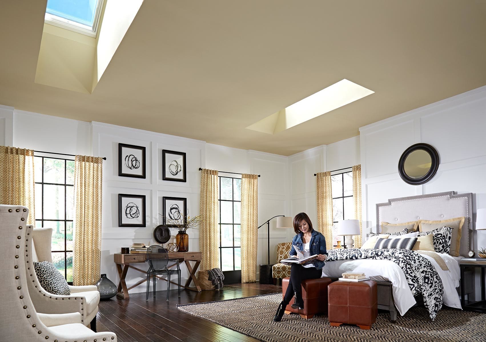 Woman sits in a bright bedroom reading a book with two skylights on the ceiling